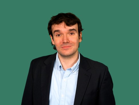 Man in black work jacket and blue collared shirt against a green background