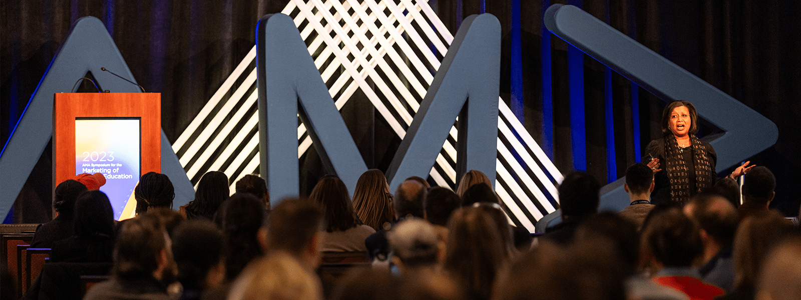 Woman on stage speaking in front of giant AMA letters at one of AMA's conferences. The audience is in the foreground and the photographer is facing the stage.